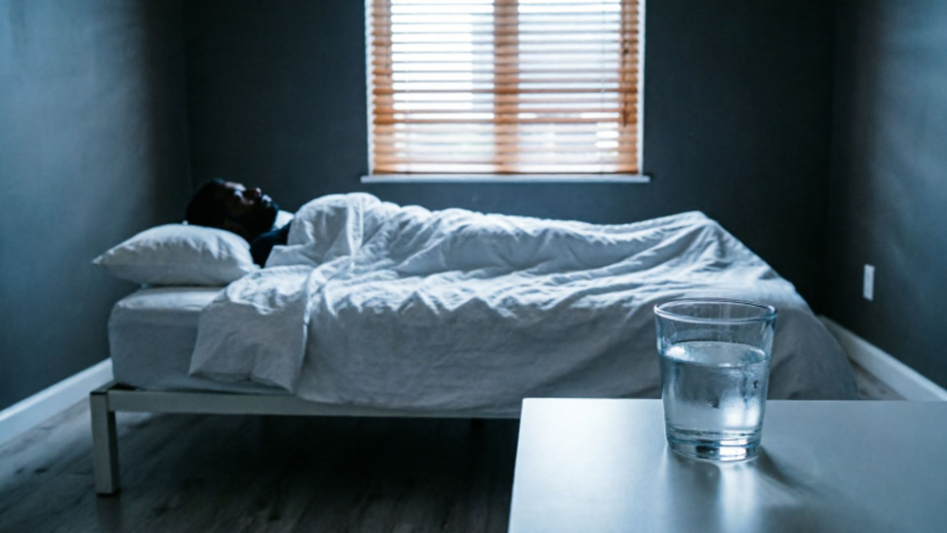 A dark minimal bedroom at pre-dawn with soft morning light filtering through blinds and a glass of water on a nightstand, representing the critical role of quality sleep in supporting healthy testosterone levels in men.
